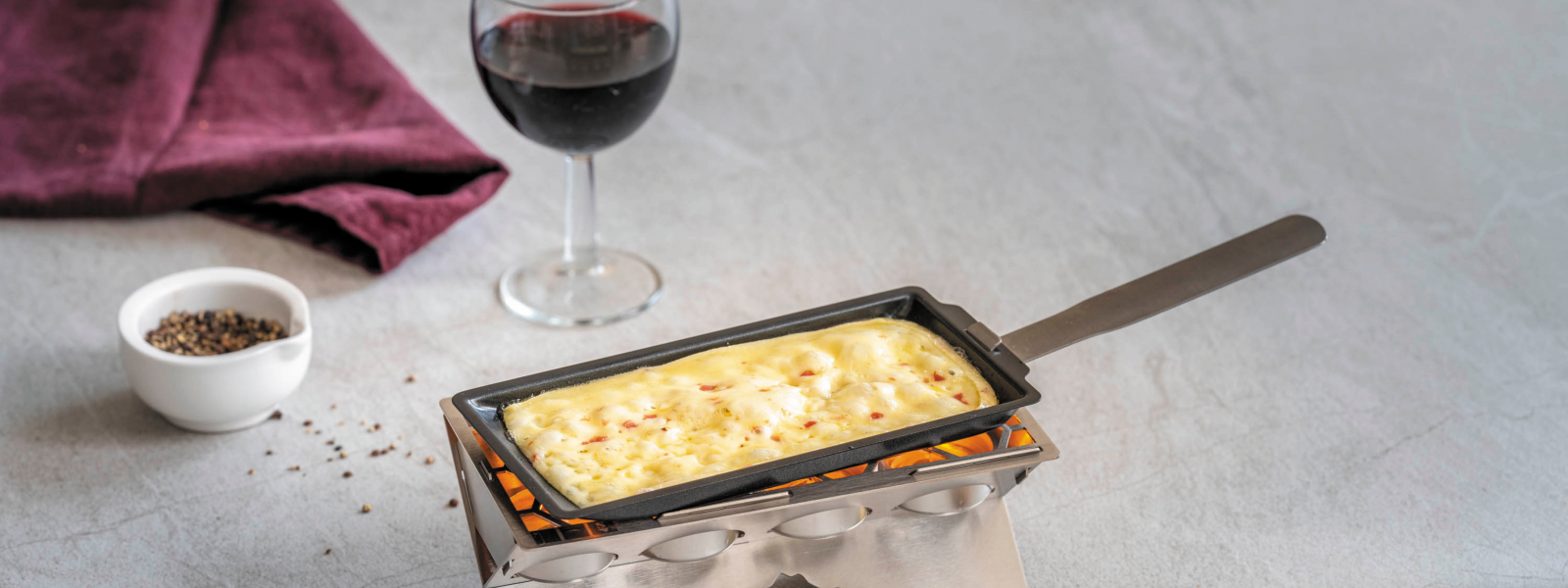 Ein Raclette Stöfchen brät Raclette, im Hintergrund ein Glas Rotwein und Pfefferkörner