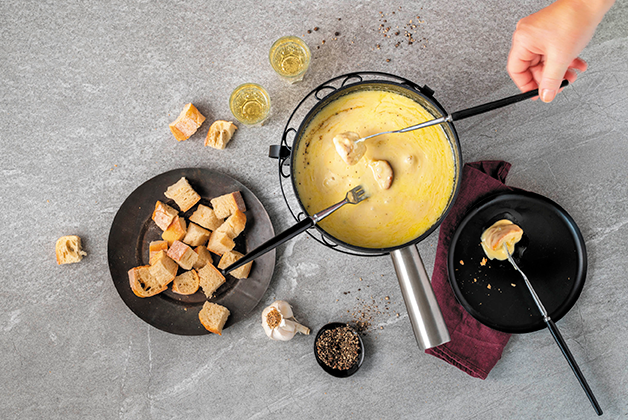 Seiler Raclette Pfannen mit geschmolzenem Käse. Ein Mann sticht mit Brot mit einer Raclette Gabel in den Käse