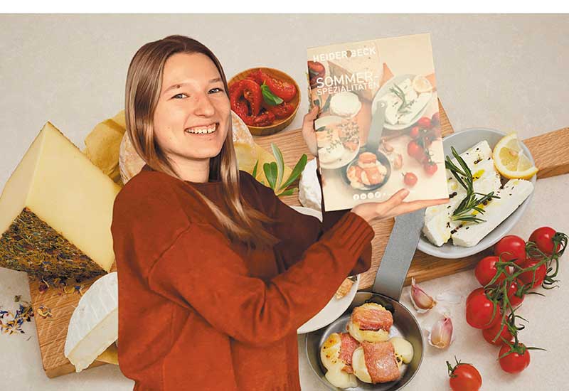 Ayleen, eine junge Dame, präsentiert den Heiderbeck Sommerkatalog 2022. Im Hintergund das Arrangement der Käseplatte, welches auch das Thema des Katalogtitelbildes ist.