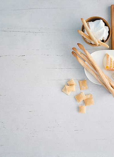 Ein Holzbrett mit verschiedenen angeschnittenen Käsen, dazu Grissini und weitere italienische Gebäckspezialitäten. Eine Schale Frischkäse rundet das Bild auf graumen Hintergrund ab