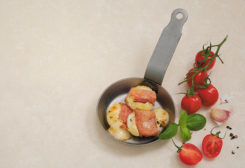 Eine Pfanne mit geschmolzenem Grillkäse aus grauem Untergrund, daneben ein Strauch Tomaten und Pfefferkörner