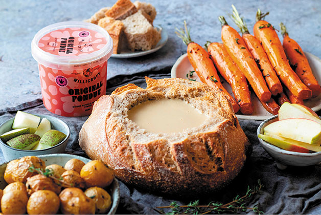 Ein ausgehöhlter Laib Brot mit veganem Fondue gefüllt, daneben eine Packung Willcroft veganes Fondue und ein Teller Möhren und ein Teller Kartoffeln