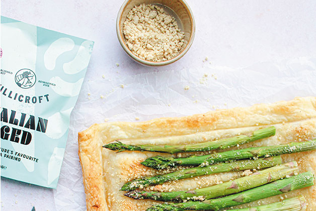 Ein Holzbrett mit grünen Spargel Stangen, die mit dem veganen Italian Aged Käse bestreut sind, daneben eine Packung Italian Aged und eine kleine Schüssel mit dem geriebenen veganen Käse.