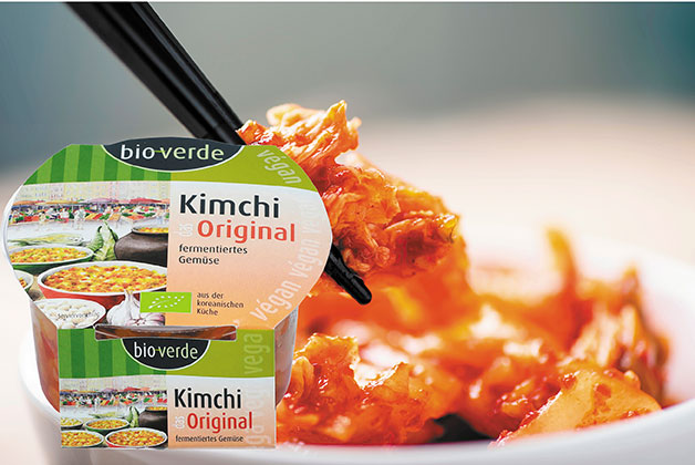 Veganes Kimchi auf einem Teller, darüber greifen zwei Stäbchen nach einem Stück Kimchi. Davor die Packung Kimchi von Bio Verde