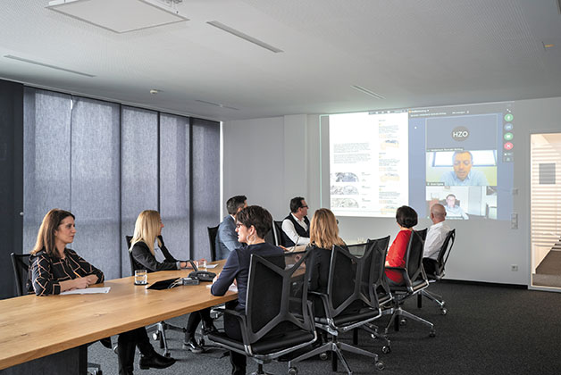 Eine Gruppe von Personen sitzen in einem Besprechungsraum bei einer Online Videokonferrenz