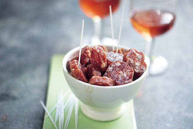 Eine kleine Schüssel mit geschnittenen Salami Stückchen auf einem Holzbrett, daneben zwei Gläser Rosé Wein